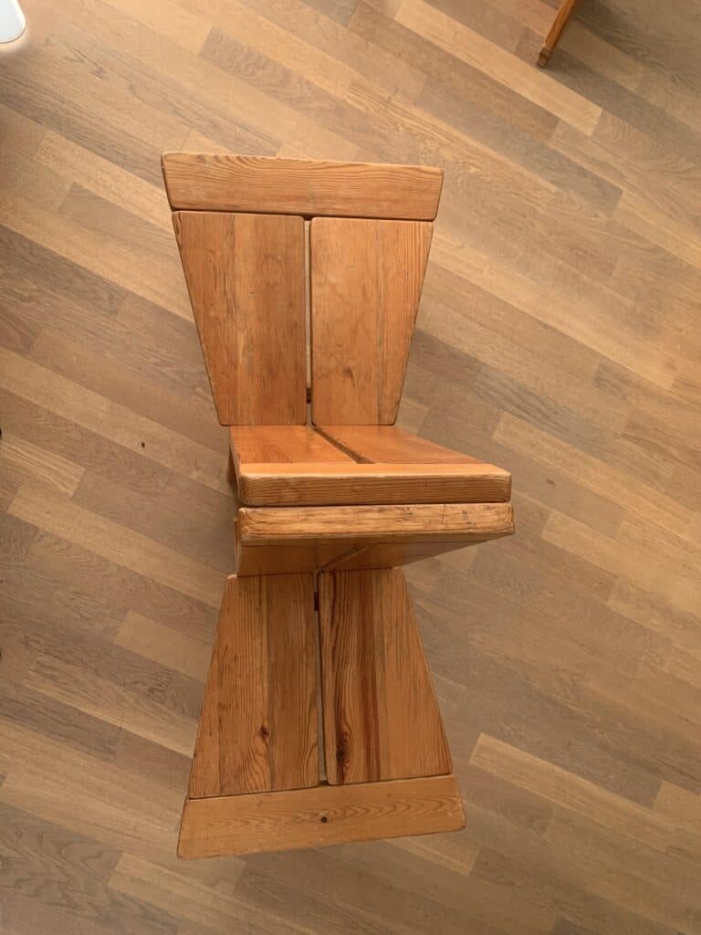 Een houten stoel met een uniek geometrisch ontwerp, met brede, platte panelen voor de zitting, rugleuning en voet, staat op een lichtbruine houten vloer.