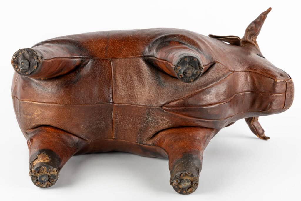 Een bruine leren poef in de vorm van een varken ligt op de rug, met vier poten met gedetailleerde hoeven en zichtbare stiksels langs het lichaam.