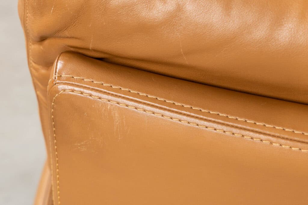 Close-up van bruine leren bekleding met zichtbare stiksels en wat lichte slijtplekken op het oppervlak.
