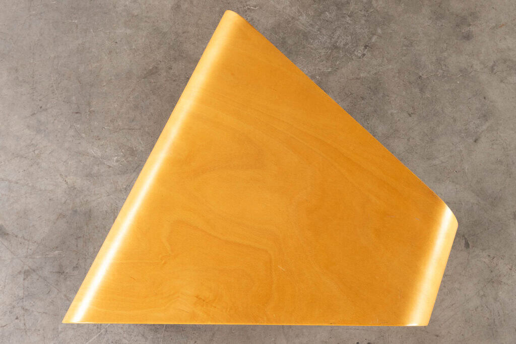 Een driehoekig stuk lichtgekleurd hout met gladde, ronde randen rust op een grijs, getextureerd betonnen oppervlak.