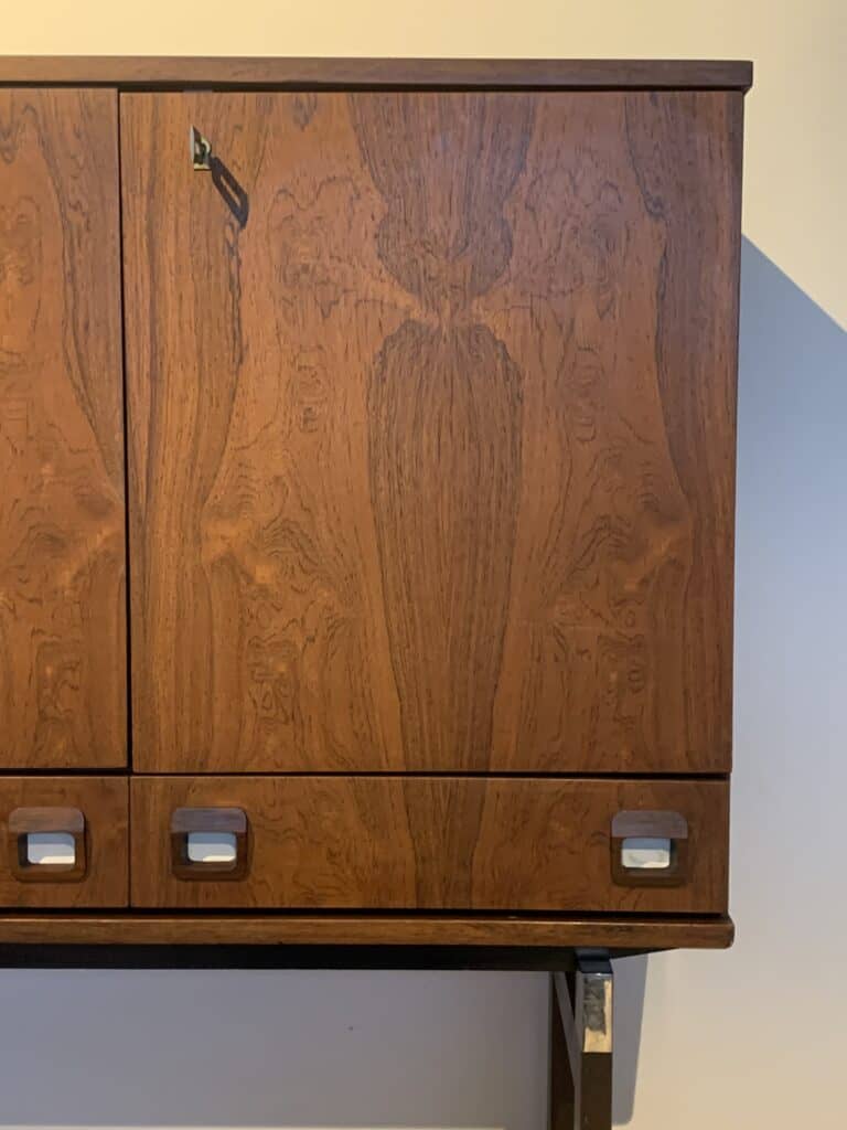 Een houten kast met ingewikkelde nerfpatronen, twee gesloten deuren, een zichtbaar slot met sleutel en drie horizontale laden aan de onderkant, elk met een rechthoekige metalen handgreep.
