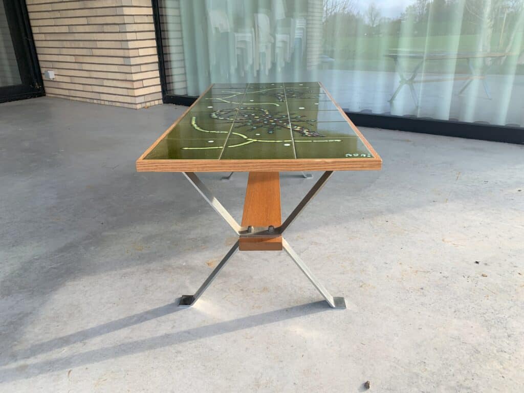 Een rechthoekige tafel met een groen, gebloemd glazen blad, houten randen en metalen poten staat op een betonnen terras voor grote glazen deuren en een bakstenen muur.