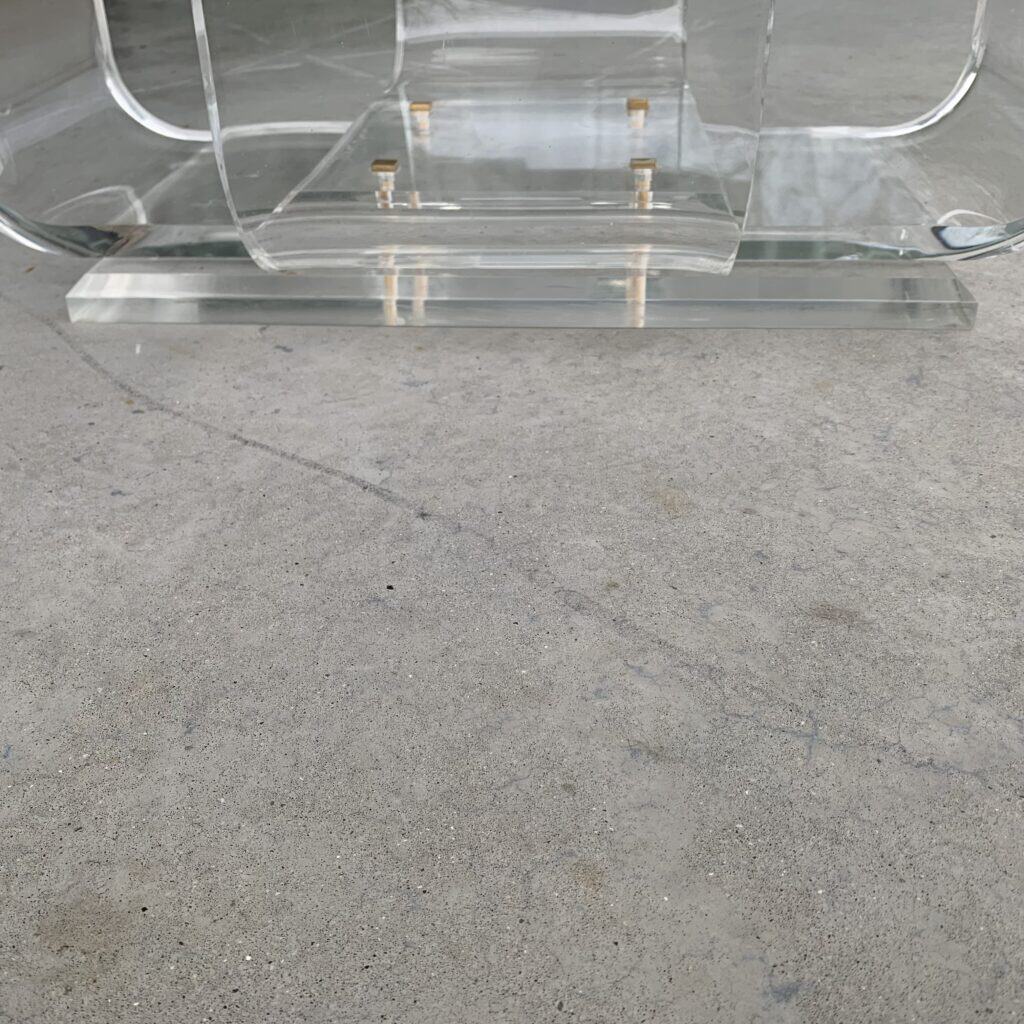 Een close-up van een stoel van helder acryl of glas met gouden details, geplaatst op een lichtgrijze betonnen vloer. De stoel en het onderstel zijn transparant en weerspiegelen een deel van de omgeving.