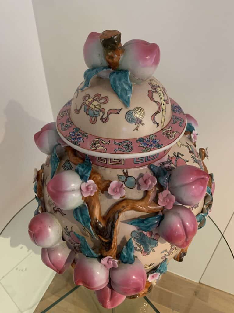 Een decoratieve keramische pot met deksel, versierd met roze en witte perziken, groene bladeren en bruine takken, met kleurrijk geschilderde motieven en patronen op een lichte achtergrond. De pot staat op een glazen oppervlak.