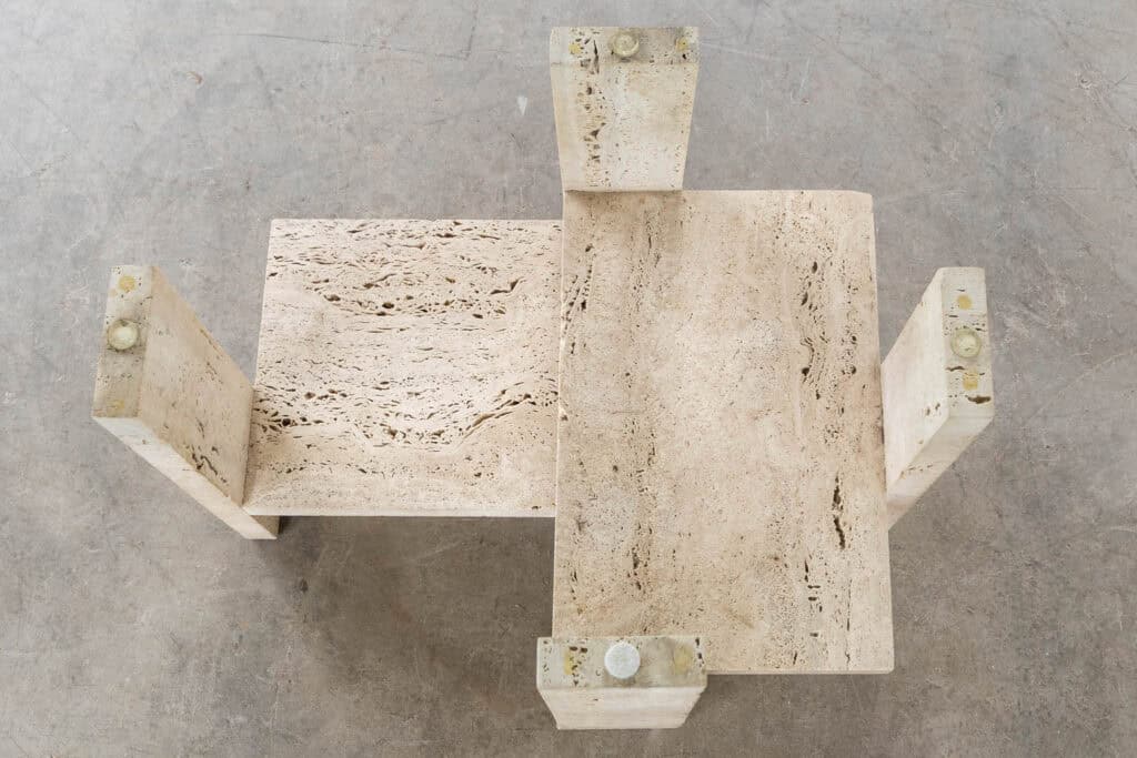 Een geometrische salontafel van beige travertijn met twee in elkaar grijpende rechthoekige vlakken en vier stevige, getextureerde poten, van bovenaf gezien op een betonnen vloer.