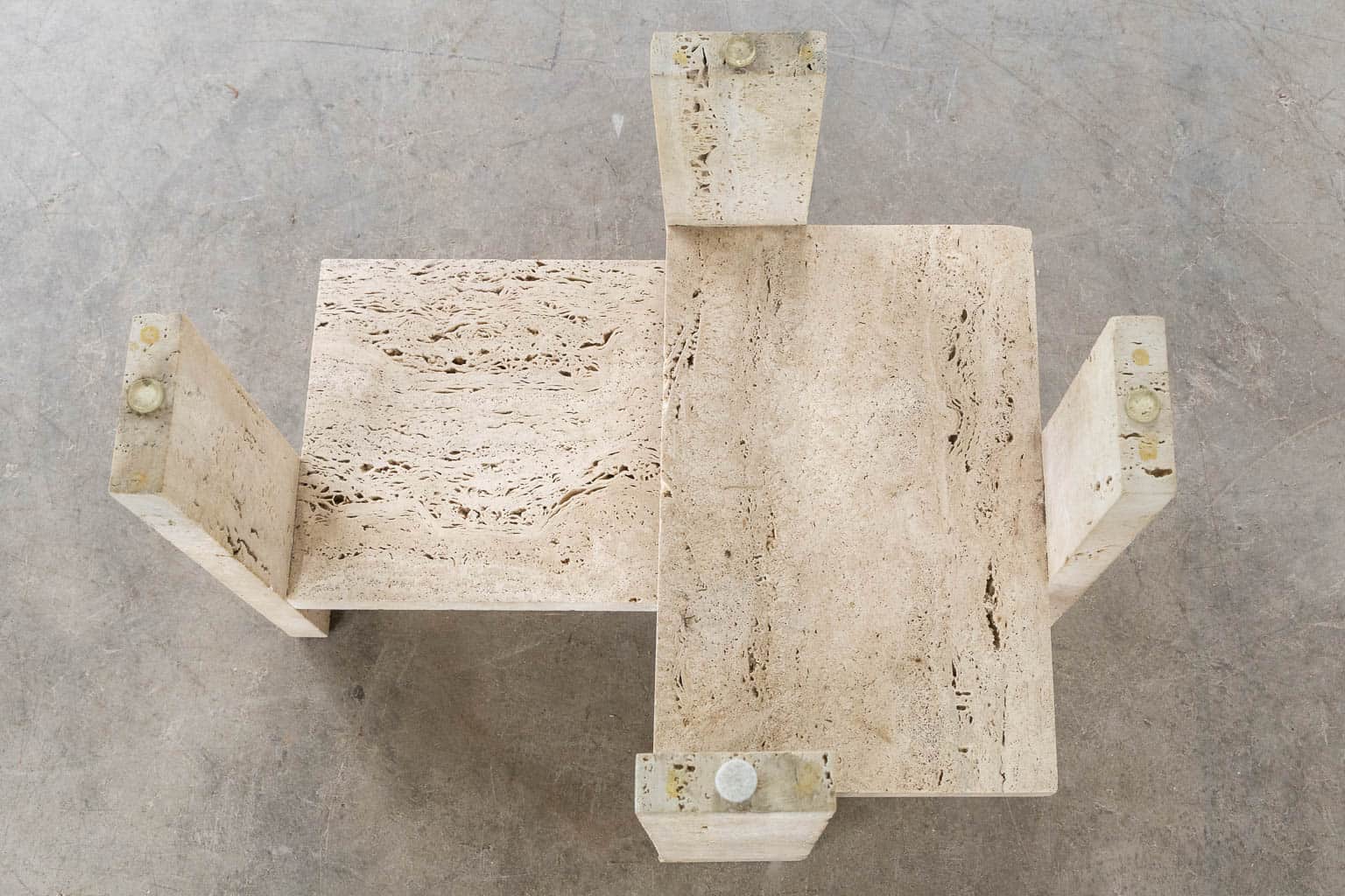 Een geometrische salontafel van beige travertijn met twee in elkaar grijpende rechthoekige vlakken en vier stevige, getextureerde poten, van bovenaf gezien op een betonnen vloer.