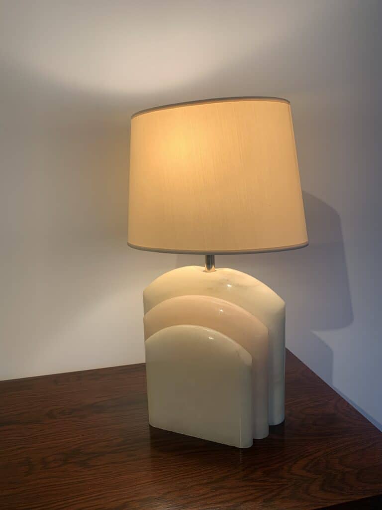 Een moderne tafellamp met een beige cilindrische kap en een glanzende, sculpturale witte keramische voet staat op een donker houten oppervlak tegen een lichtgekleurde muur. De lamp is aan en werpt een warm licht.