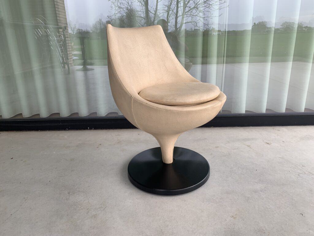 Een moderne beige draaistoel met een gebogen rugleuning en een rond zwart onderstel staat op een betonnen vloer voor glazen deuren met doorschijnende witte gordijnen. Buiten zijn bomen en gras zichtbaar.