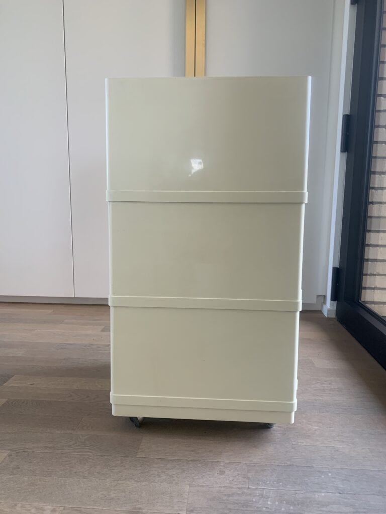 Een rechthoekige, gebroken witte luchtreiniger met wielen staat op een houten vloer naast een glazen deur en een witte muur.