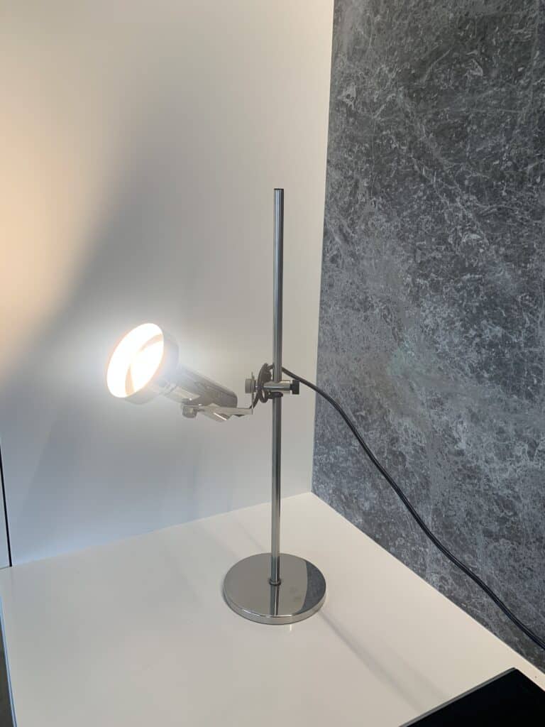 Een moderne, verstelbare metalen bureaulamp met een ronde voet en een spotlampkop verlicht een wit oppervlak naast een grijs gemarmerde muur. Er hangt een zwart netsnoer aan de lamp.