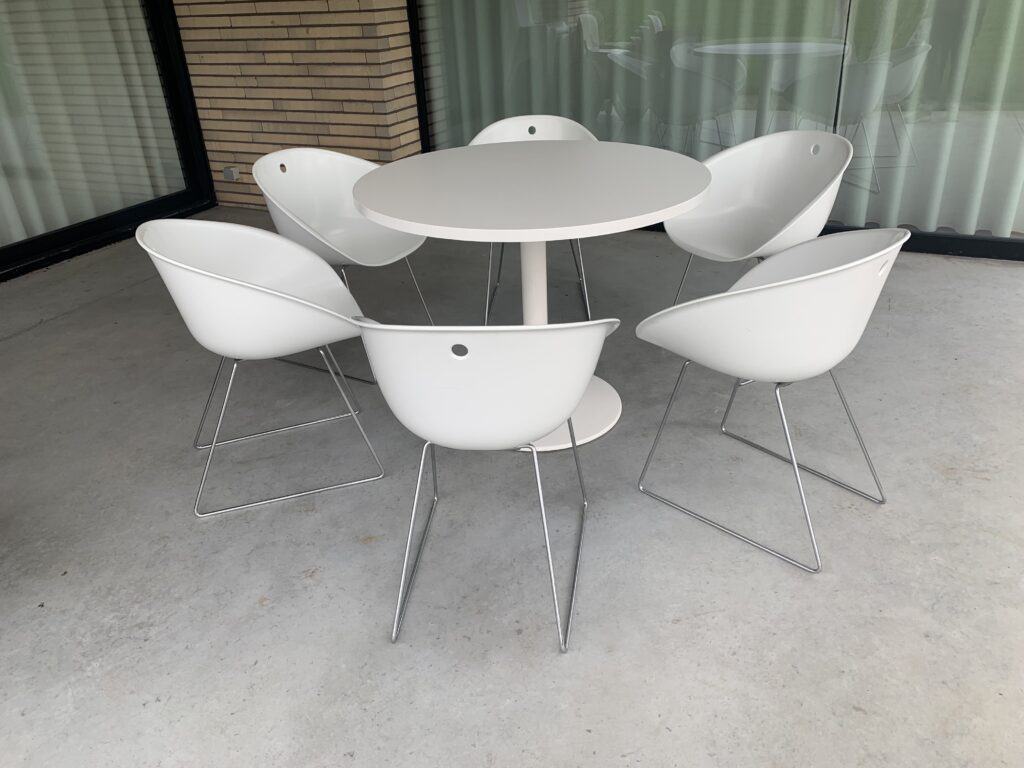 Een ronde witte tafel met daaromheen zes moderne witte stoelen op metalen poten, op een terras van beton met grote ramen met gordijnen en een bakstenen muur.
