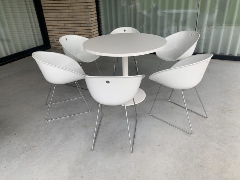 Een ronde witte tafel met daaromheen zes moderne witte stoelen op metalen poten, op een terras van beton met grote ramen met gordijnen en een bakstenen muur.