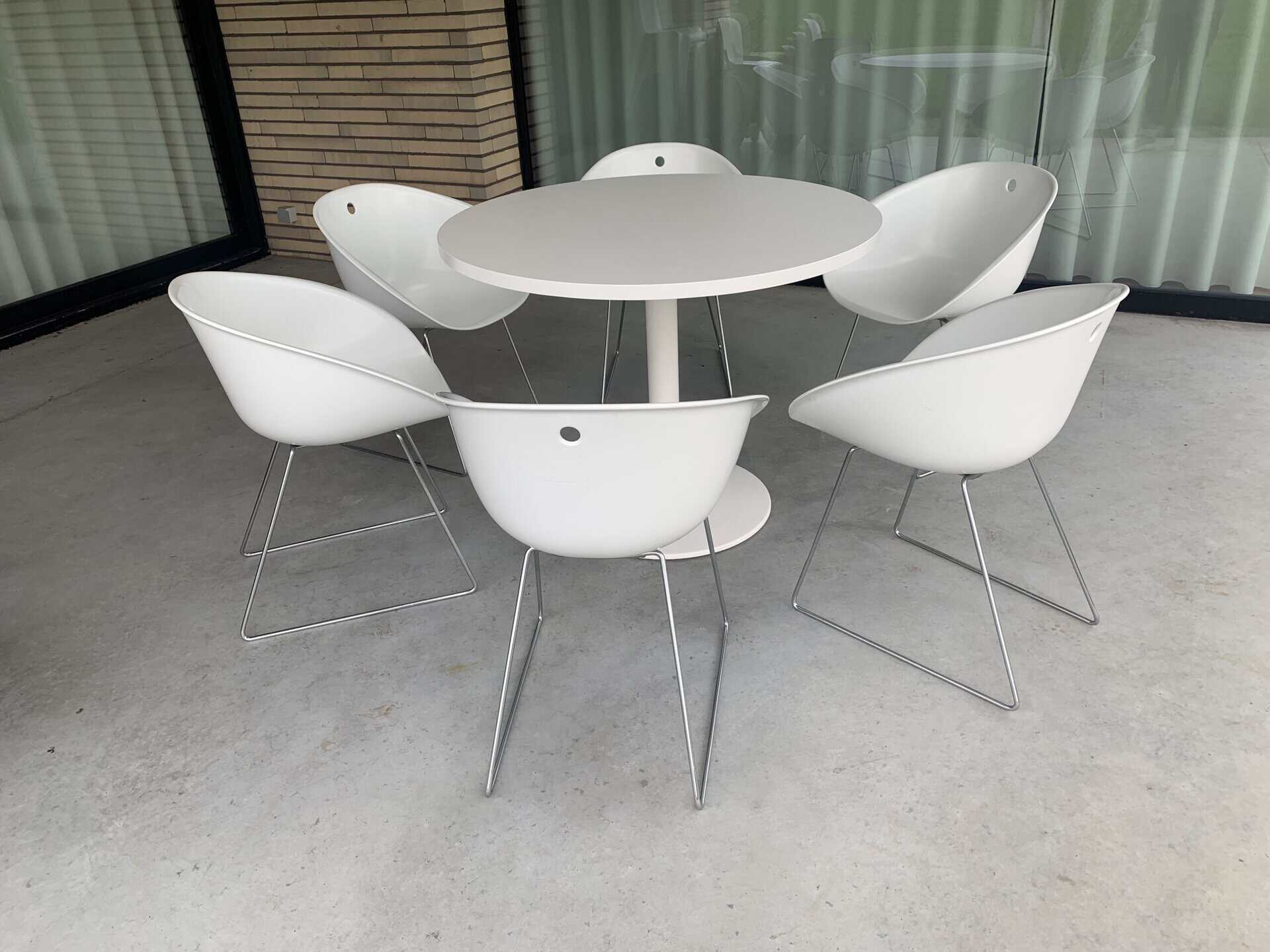 Een ronde witte tafel met daaromheen zes moderne witte stoelen op metalen poten, op een terras van beton met grote ramen met gordijnen en een bakstenen muur.