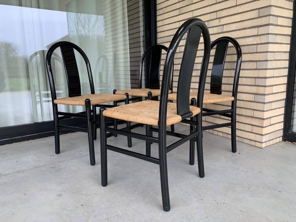 Vier zwarte houten stoelen met gevlochten rieten zittingen staan in een trapsgewijs patroon op een betonnen terras, naast een bakstenen muur en een groot raam.