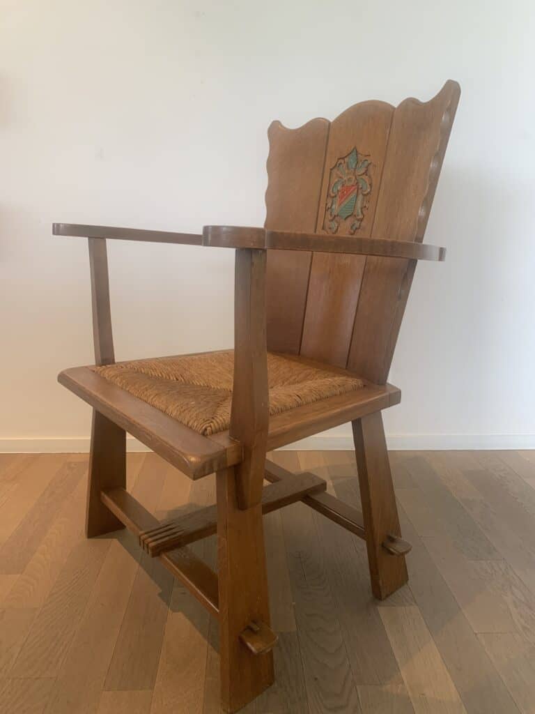 Een houten fauteuil met gesneden poten en armleuningen, een zitting van gevlochten riet en een decoratief bloemenmotief op de rugleuning, geplaatst op een lichte houten vloer tegen een eenvoudige witte muur.