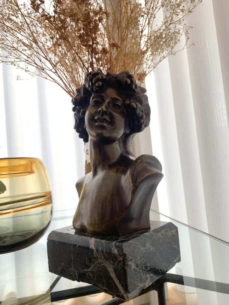 Een bronzen buste van een vrouw met krullend haar staat op een donkere marmeren voet boven op een glazen tafel; gedroogde bloemen en zonlicht stromen door dunne gordijnen op de achtergrond.