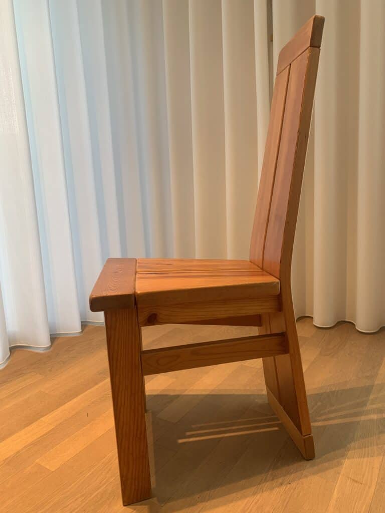 Een houten stoel met een rechte rugleuning en een eenvoudig ontwerp, geplaatst op een lichte houten vloer voor witte gordijnen, gefotografeerd vanaf de zijkant.