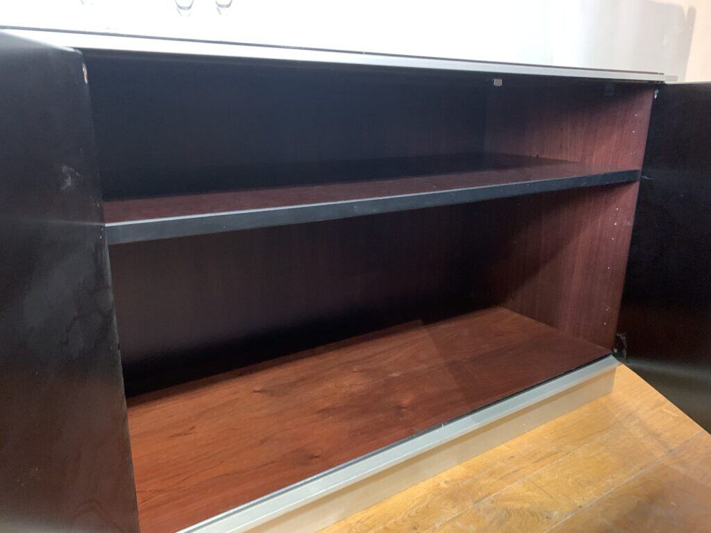 Een lege, donkere houten kast met twee openstaande deuren en een enkele horizontale plank erin, staand op een lichte houten vloer. De binnenkant van de kast is schoon en leeg.