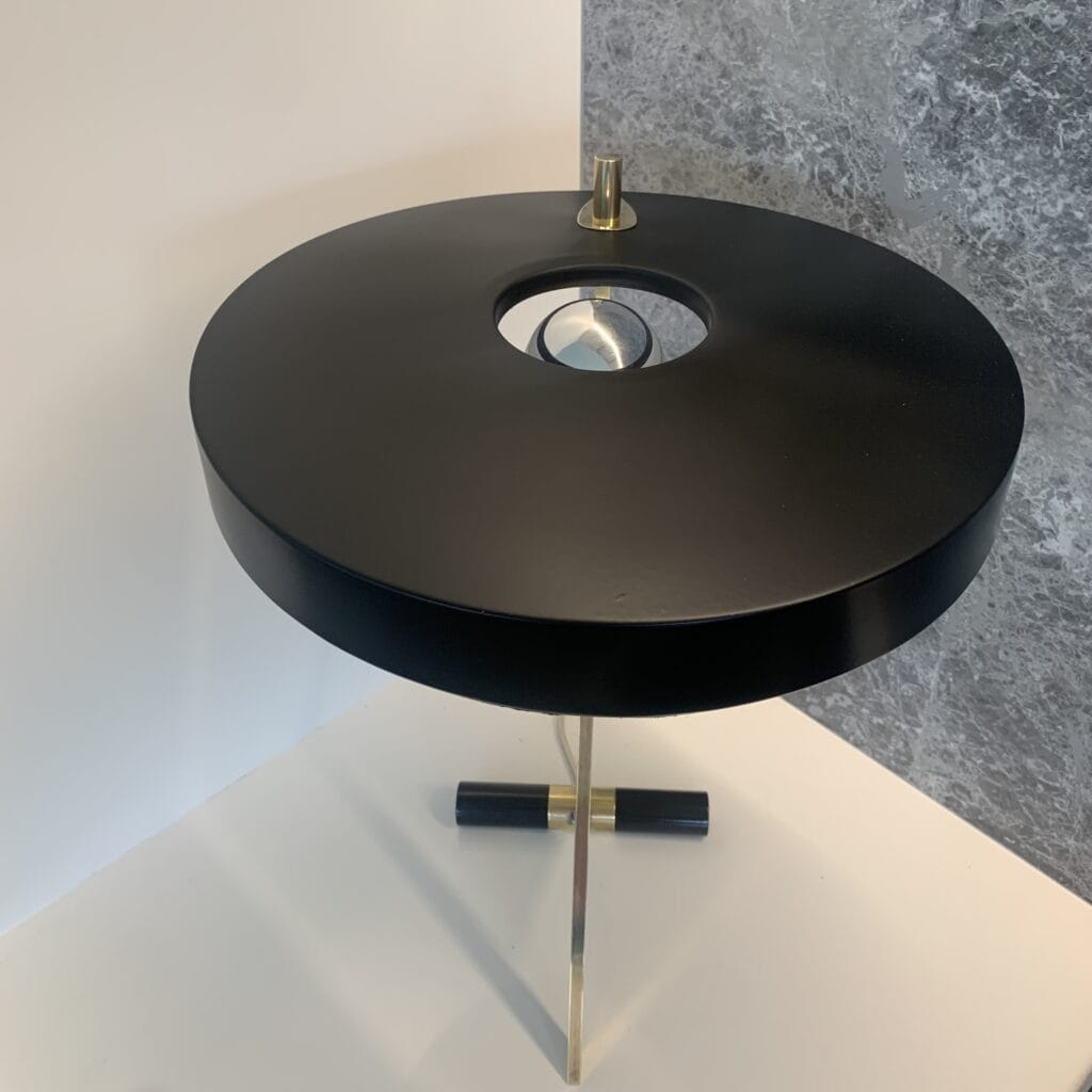 Een moderne, ronde, zwarte bijzettafel met een messing onderstel en een ronde uitsparing in het midden, geplaatst tegen een tweekleurige muur met witte en grijze structuuroppervlakken.