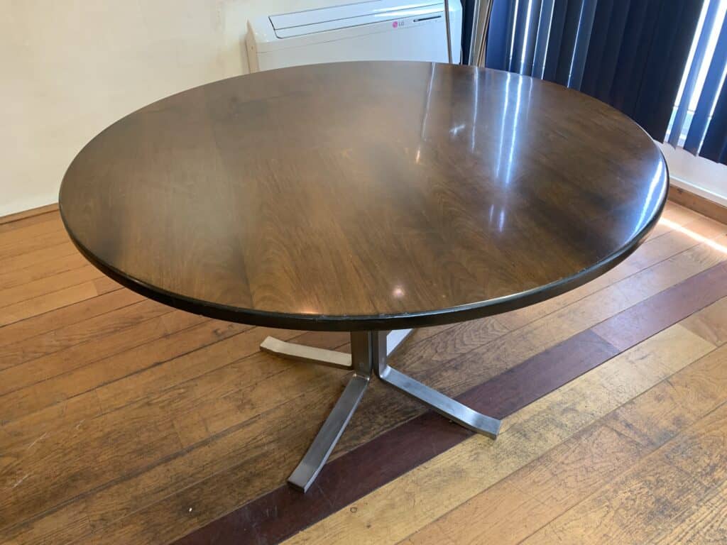 Een ronde houten tafel met een glanzend oppervlak en een metalen voetstuk staat op een hardhouten vloer, vlak bij een aan de muur gemonteerde airconditioning en donkere verticale jaloezieën.