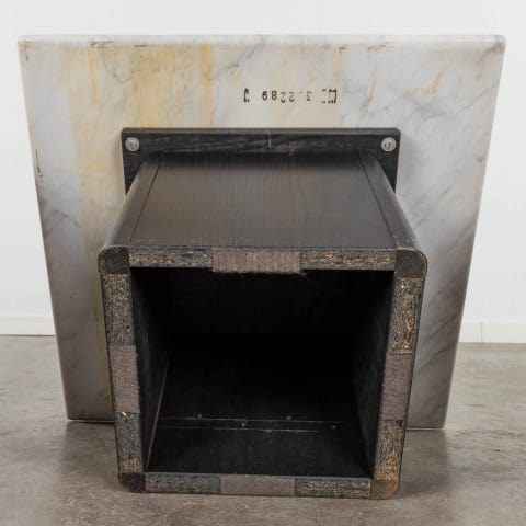 Een vierkante, zwarte, holle sculptuur met getextureerde randen is gemonteerd op een marmeren plaat, rechtopstaand op een betonnen vloer tegen een witte muur. Een serienummer is zichtbaar op het marmeren oppervlak.
