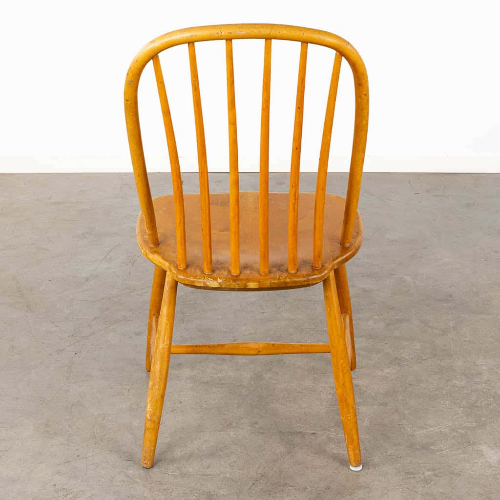 Een houten stoel met een gebogen rugleuning en verticale spijlen staat, van achteren gezien, op een grijze betonnen vloer tegen een eenvoudige witte muur.