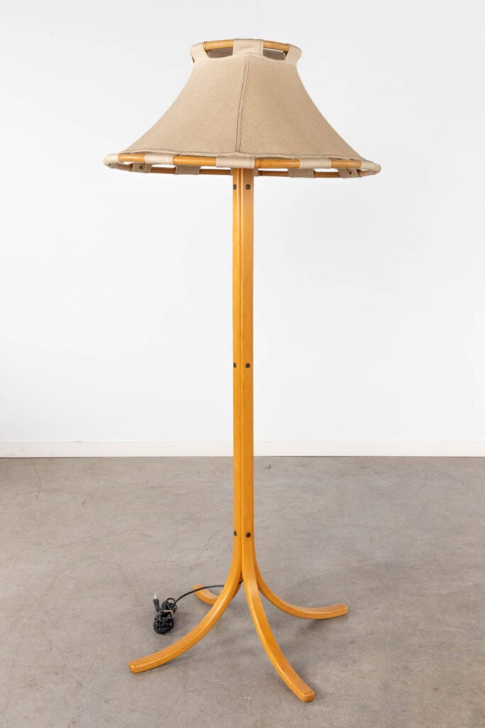 Een houten staande lamp met een beige stoffen kap, staand op vier gebogen poten, staat op een grijze betonnen vloer tegen een effen witte muur. Het zwarte snoer van de lamp is zichtbaar op de vloer.