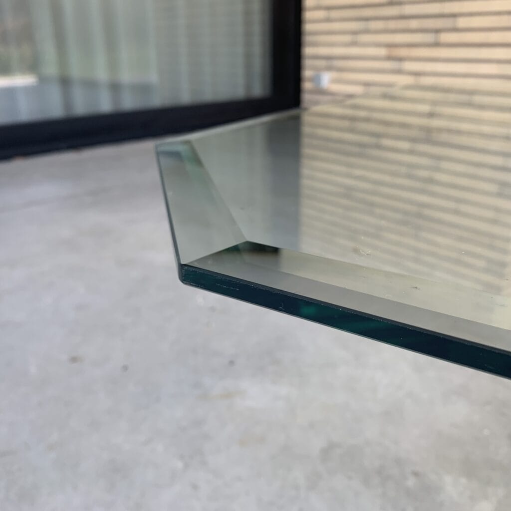Close-up van de scherpe hoek van een glazen tafel of paneel, met een onscherpe achtergrond waarop een betonnen vloer, een raam met een zwart kozijn en een bakstenen muur te zien zijn.
