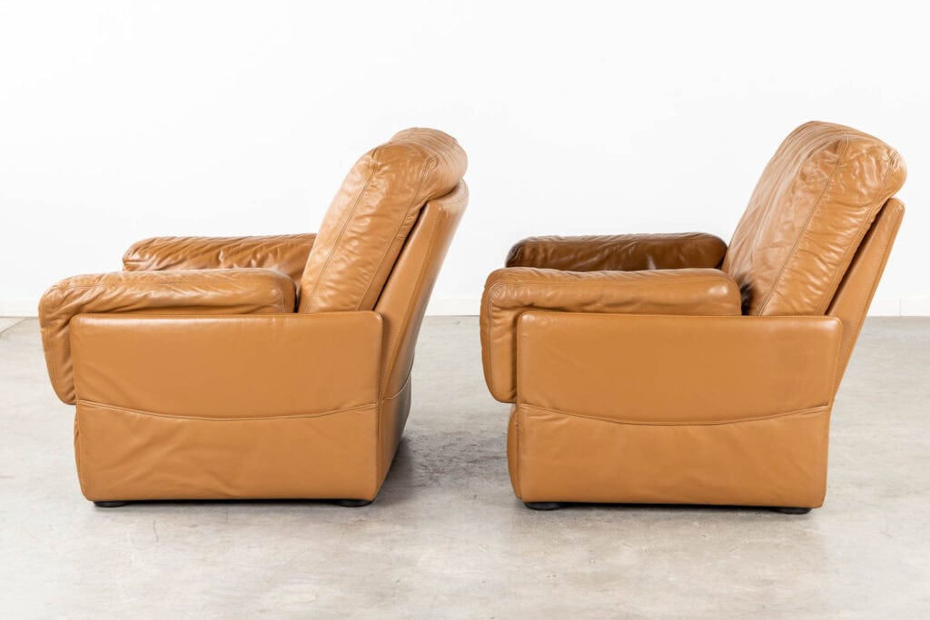 Twee bruine leren fauteuils staan naast elkaar, van opzij gezien tegen een effen witte muur en een grijze betonnen vloer. De stoelen hebben dikke, zachte rugleuningen, armleuningen en zittingen.