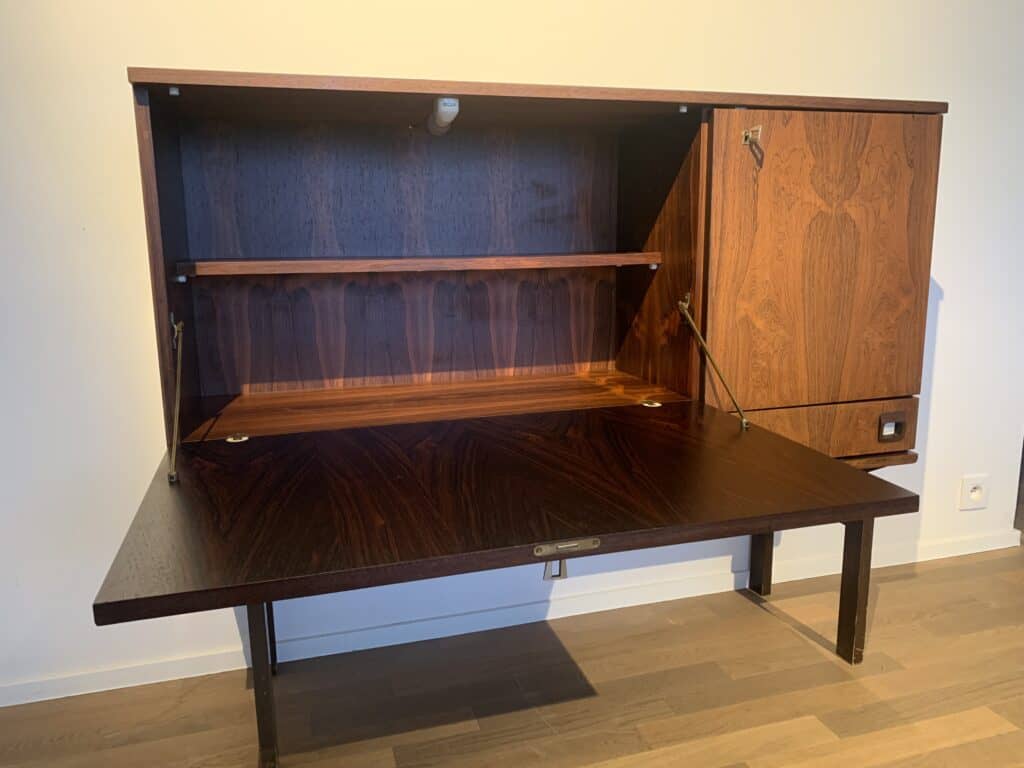 Een vintage houten wandbureau met een open, uitklapbaar schrijfoppervlak, planken aan de binnenkant en een klein kastdeurtje aan de rechterkant, tegen een lichtgekleurde muur en houten vloer.