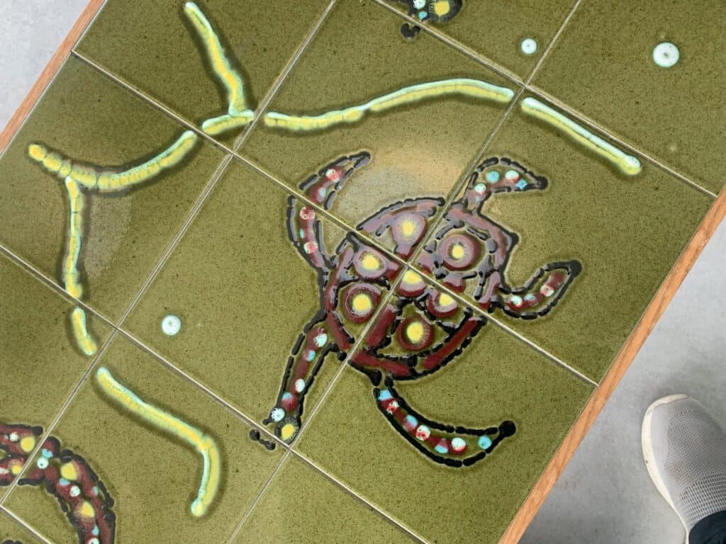 Een close-up van een betegeld tafelblad met een kleurrijk, abstract schildpaddessin met stippen op een groene achtergrond. Rechtsonder is een deel van een witte sneaker zichtbaar.