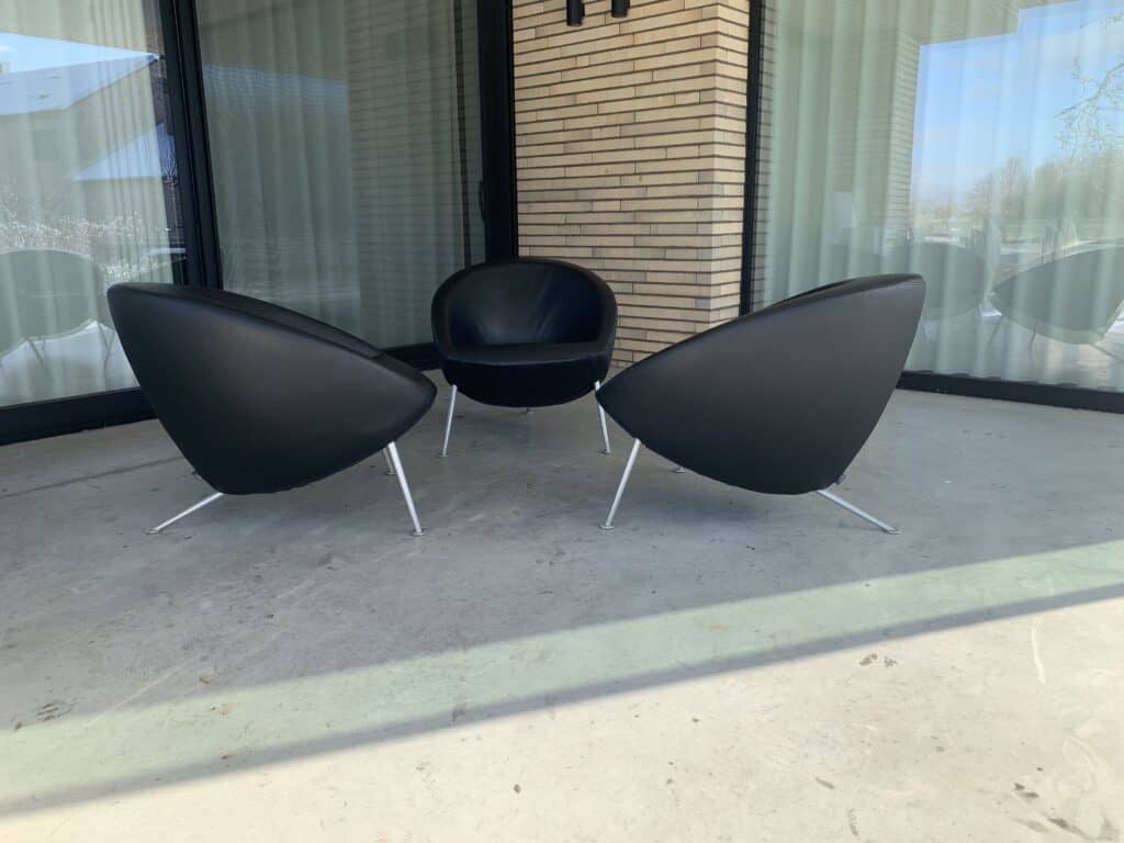 Drie moderne, zwarte, ronde stoelen met metalen poten staan in een cirkel op een betonnen terras buiten een gebouw met bakstenen muren, grote ramen en doorschijnende witte gordijnen.