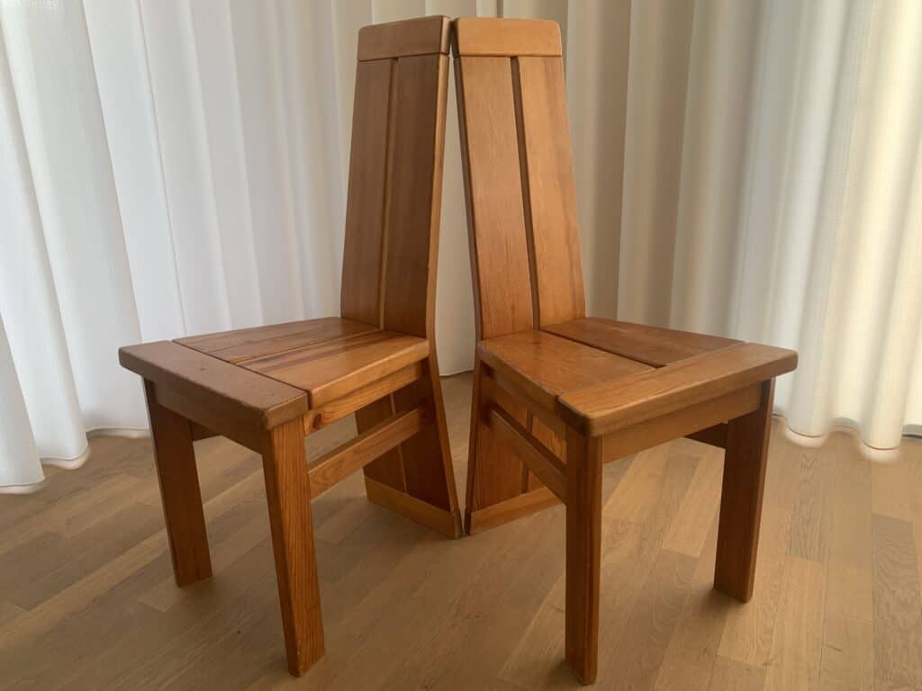 Twee houten stoelen staan rug aan rug met de bovenhoeken tegen elkaar, waardoor een gespiegelde, symmetrische opstelling ontstaat. De stoelen hebben hoge, schuine rugleuningen en staan op een lichte houten vloer met witte gordijnen op de achtergrond.