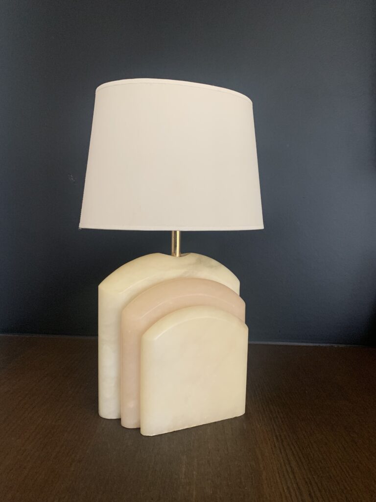 Een tafellamp met een witte kap en een voet van drie overlappende, gebogen stenen stukken in crème- en lichtroze tinten, op een donker houten oppervlak tegen een zwarte muur.