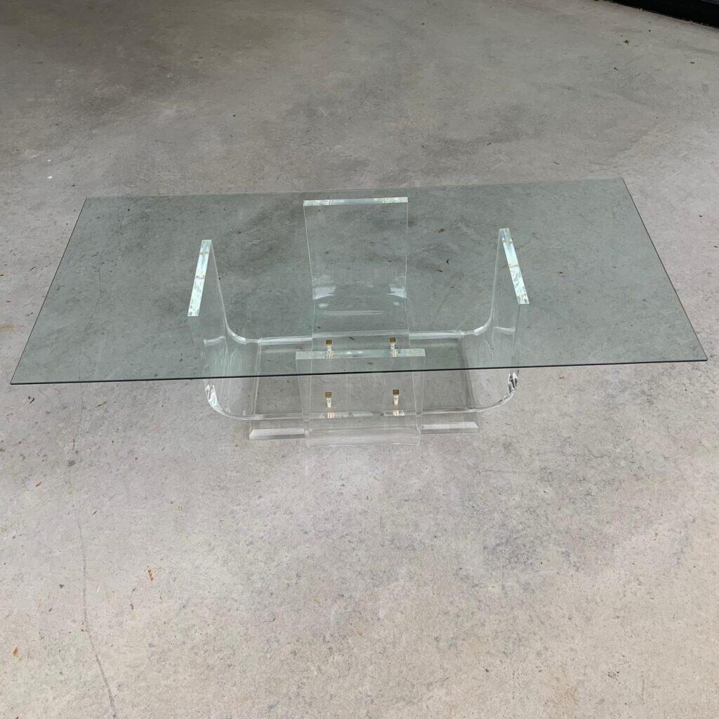 Een rechthoekige glazen salontafel met een transparant acryl onderstel, geplaatst op een lichtgekleurde betonnen vloer. Het ontwerp kenmerkt zich door strakke lijnen en een moderne esthetiek.