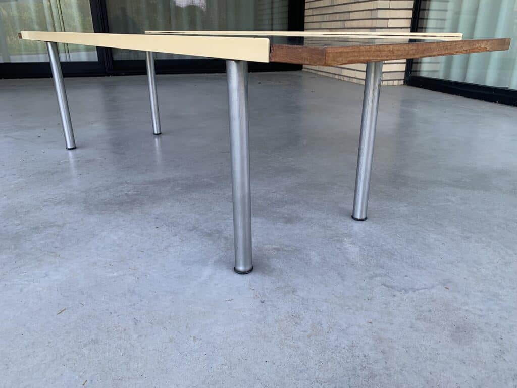 Een tafel met vier metalen poten op een grijs betonnen terras. Het tafelblad is ongelijk, met één kant hoger en lichtgekleurd, en de andere kant lager en van donker hout. Glazen deuren en een bakstenen muur zijn op de achtergrond te zien.