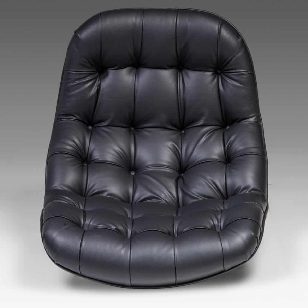 Een moderne, zwarte leren loungestoel met dikke, gewatteerde kussens en een gebogen, armloos ontwerp, tegen een effen grijze achtergrond.