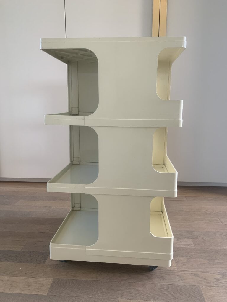Een beige, drielaags plastic opbergkarretje met open zijkanten staat op een houten vloer in een lichte kamer. Het karretje heeft wielen en is leeg, waardoor de rechthoekige planken en het eenvoudige ontwerp goed te zien zijn.