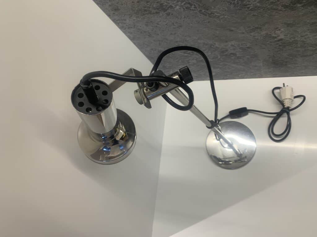 Een metalen bureaulamp met verstelbare arm en klem staat op een wit oppervlak, naast een opgerolde zwarte stroomkabel met een witte stekker. De achtergrond bestaat uit een grijze muur met textuur.