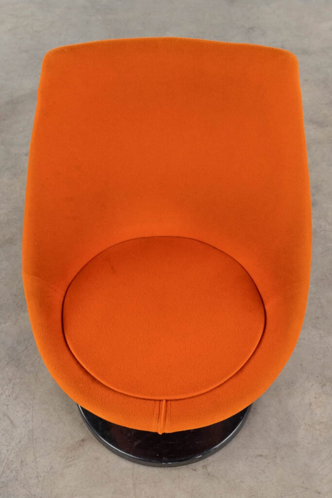 Een oranje, moderne, gestoffeerde stoel met een ronde rugleuning en zitting, van bovenaf gezien. De stoel staat op een rond metalen onderstel, geplaatst op een grijze betonnen vloer.