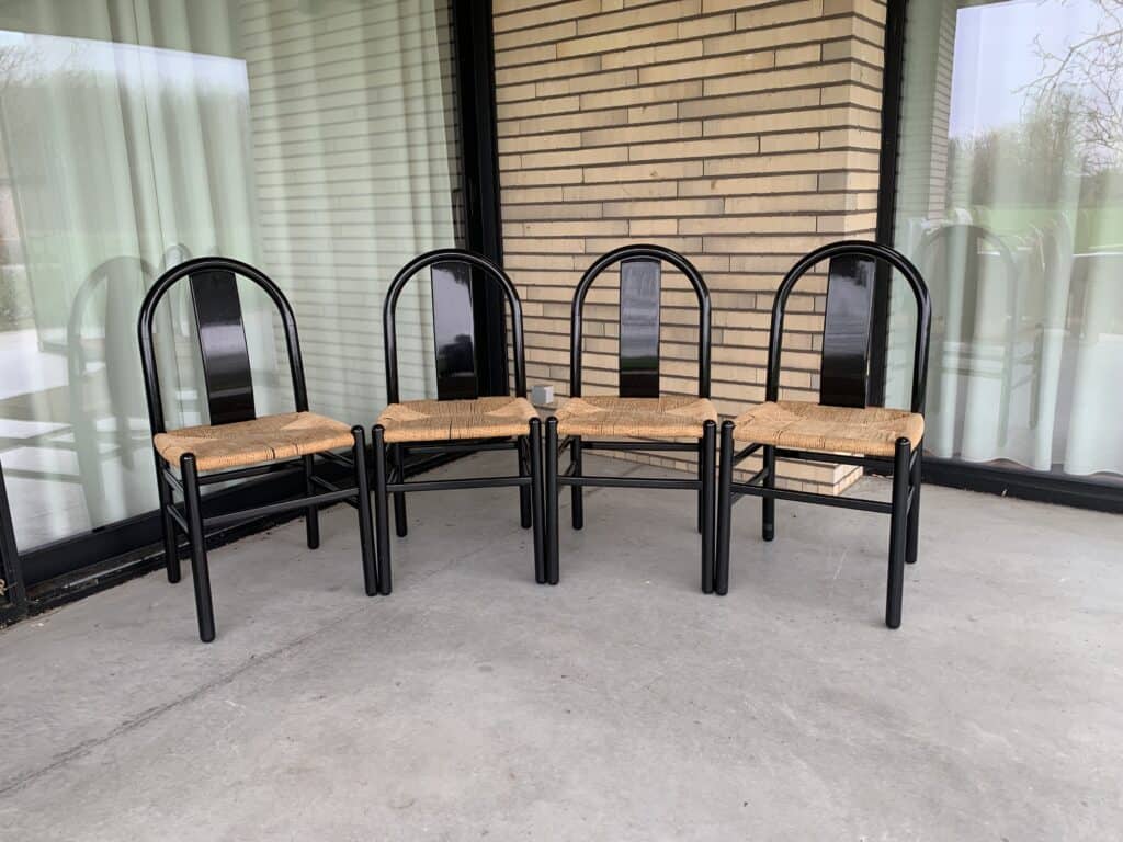 Vier zwarte metalen stoelen met gevlochten zittingen staan in een halve cirkel op een grijs betonnen terras, vlakbij een glazen raam en een bakstenen muur met zichtbare gordijnen.