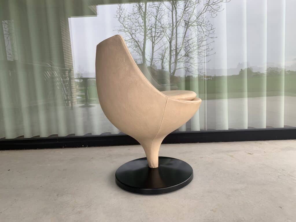 Een moderne, beige tulpvormige stoel met een ronde zitting en rugleuning staat op een zwarte ronde voet. De stoel staat op een betonnen vloer voor grote glazen schuifdeuren met transparante gordijnen.