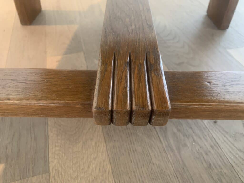 Close-up van twee elkaar kruisende houten balken met een gesneden verbinding die lijkt op vier vingers die één balk vasthouden, op een lichte houten vloer.