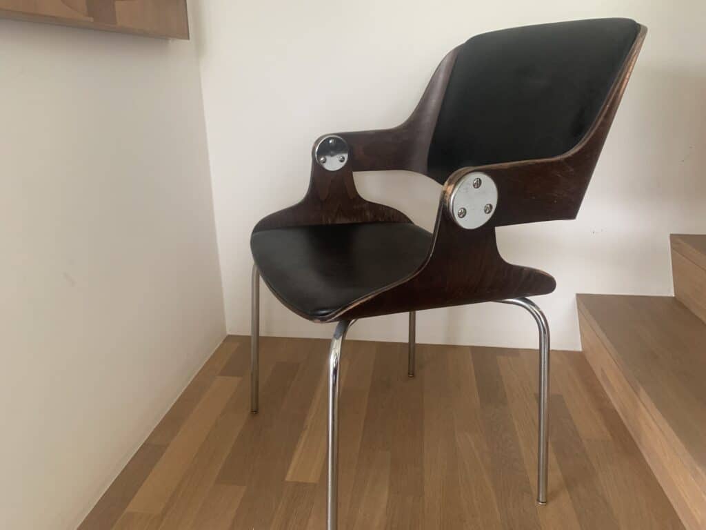 Een moderne stoel met een donker houten frame, een zwarte, gevoerde zitting en rugleuning, metalen armleuningen en chromen poten, geplaatst op een houten vloer vlak bij treden en een witte muur.