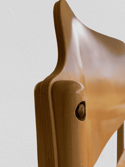 Close-up van de rugleuning en het bovenste deel van een houten stoel met een zichtbare schroef, tegen een eenvoudige, lichte achtergrond.