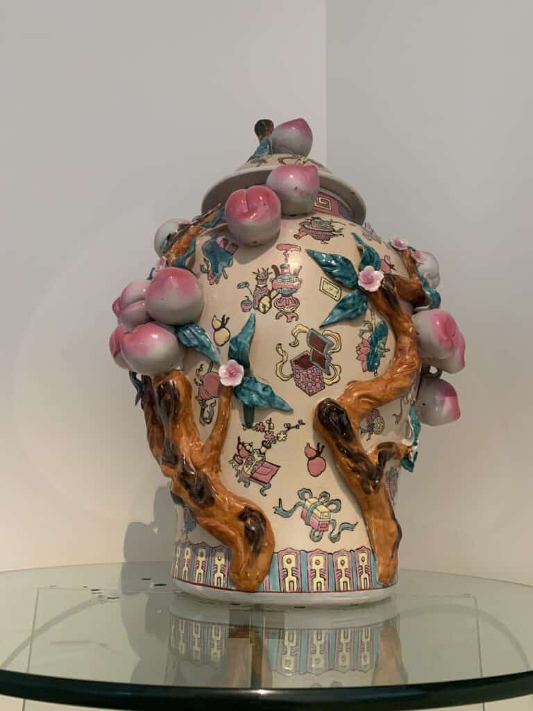 Een decoratieve keramische pot met deksel, waarop symbolen en ingewikkelde ontwerpen zijn geschilderd, versierd met gebeeldhouwde takken, roze perziken en groene bladeren. De pot staat op een glazen plank tegen een witte achtergrond.