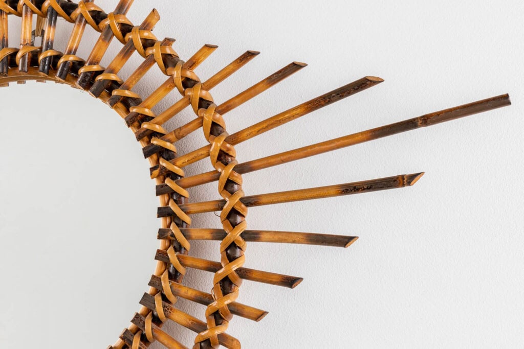 Close-up van een decoratieve ronde spiegel met een ingewikkeld gevlochten bamboe frame, met stralende spaken in de vorm van zonnestralen tegen een witte muur.