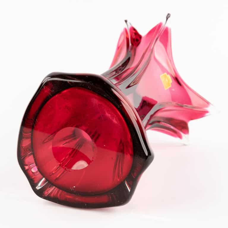 Een rood-roze glazen vaas met een uitlopende, stervormige rand ligt op zijn kant tegen een witte achtergrond. De voet is rond en dieprood, met decoratieve krullen en een klein gouden label.