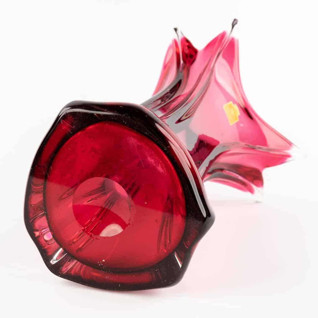 Een rood-roze glazen vaas met een uitlopende, stervormige rand ligt op zijn kant tegen een witte achtergrond. De voet is rond en dieprood, met decoratieve krullen en een klein gouden label.