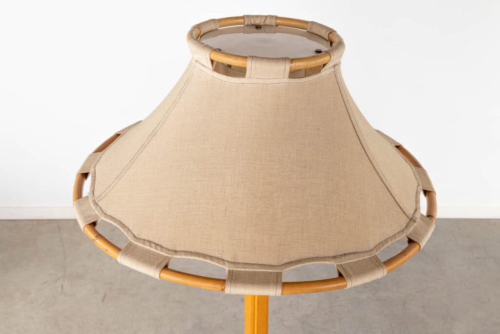 Een close-up van een beige stoffen lampenkap met een houten frame en decoratieve lussen rond de rand, tegen een lichtgekleurde muur en grijze vloer.